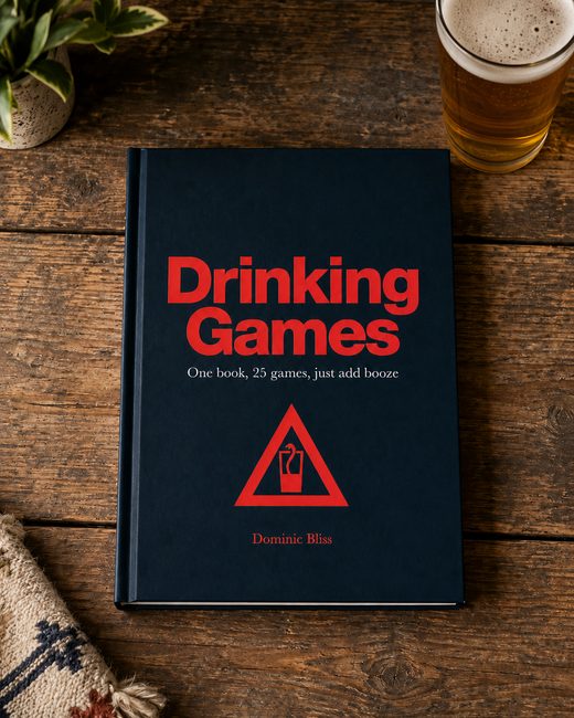 Dark blue drinking games book with red logo on a wooden table next to a glass of beer.